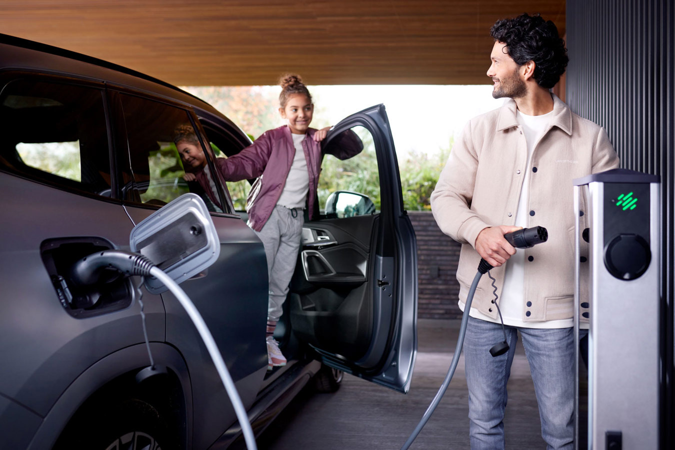 Man laadt een elektrische auto bij een hotel-laadstation terwijl zijn dochter vanuit de auto naar hem kijkt, als symbool voor zorgeloos elektrisch reizen.